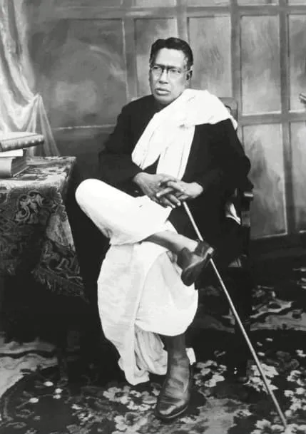 Full-length studio photograph of Bipin Chandra Pal seated in a chair, in spectacles, wearing a dark coat over a white dhoti and shawl, holding a walking stick, beside a table with books, against a painted backdrop.